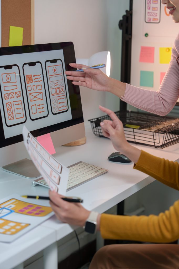 Two web designers are discussing user interface of mobile application displayed on desktop computer