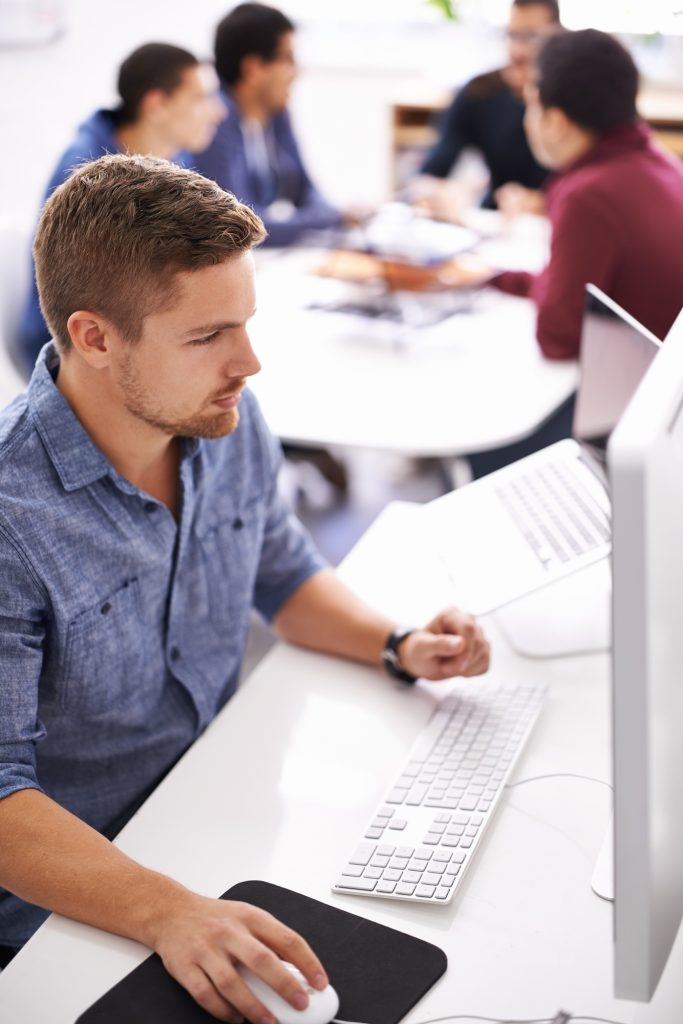 Shot of a young designer working at his computer with colleagues in the background